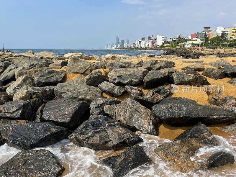 斯里兰卡科伦坡拉维尼亚山海滩上的裂缝/岩石盔甲图像，沙滩和岩石海滩，背景中可以看到大海和首都，海岸侵蚀防御设施和晴天的蓝天
